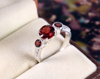 May include: A silver ring featuring three red gemstones. The center stone is larger and round, flanked by two smaller, round stones. The band is detailed with small, clear stones. The ring is presented in a brown jewelry box with a cream-colored interior.