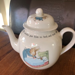 May include: White ceramic teapot with a Peter Rabbit illustration. The teapot features a quote from the book, "His mother put him to bed, and made sure..."