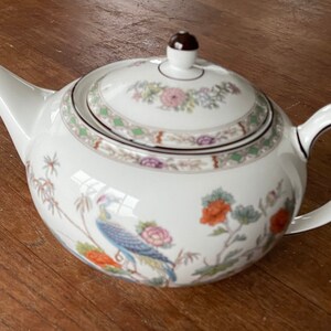 Pode incluir: Bule de porcelana branco com tampa, com um design floral e de pássaros coloridos. O bule tem um bico curvo, uma alça arredondada e um botão marrom na tampa. O design inclui flores laranja, azuis e rosas e um pavão.