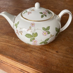Op de afbeelding: Een witte porseleinen theepot met een delicaat botanisch patroon van roze bloemen, groene bladeren en dunne bruine ranken. De theepot is voorzien van een bijpassend deksel met een kleine knop, een gebogen handvat en een tuit, allemaal afgewerkt met een dunne gouden rand.