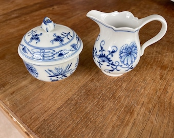 May include: A white porcelain creamer and sugar bowl set with blue floral designs. The creamer has a handle and a spout. The sugar bowl has a lid with a knob.
