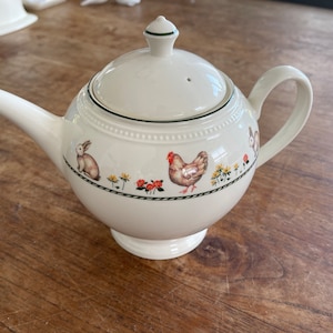 Könnte beinhalten: Cremefarbene Keramik-Teekanne mit Deckel und Henkel. Die Teekanne ist mit einem Rand aus Hasen, Hühnern, Blumen und Blättern verziert. Der Deckel hat einen kleinen grünen Knopf und ein Entlüftungsloch. Die Teekanne steht auf einem kleinen Sockel.