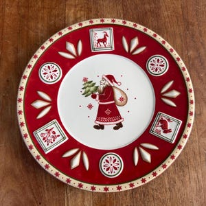 Puede incluir: Un plato redondo de cerámica con un centro blanco que muestra una ilustración de Santa Claus cargando un árbol de Navidad y un saco. El borde rojo ancho está decorado con relieves blancos en forma de hoja, motivos florales y paneles cuadrados.