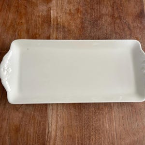 Puede incluir: Una bandeja rectangular de color blanco roto con detalles florales decorativos en las asas. La bandeja de cerámica está colocada sobre un fondo de madera. La bandeja mide aproximadamente 30 cm de largo.