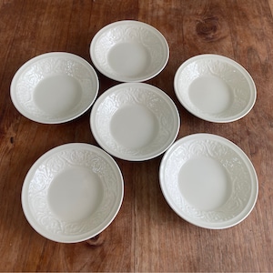 Può includere: Sei ciotole in ceramica bianca con un motivo decorativo in rilievo. Le ciotole sono disposte in cerchio su una superficie di legno.