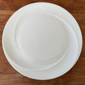 Könnte beinhalten: Ein weißer Keramikteller mit einem einzigartigen, wirbelnden Design in der Mitte. Der Teller hat einen breiten Rand und eine glatte, glänzende Oberfläche. Der Teller steht auf einer Holzoberfläche. Geeignet zum Servieren von Speisen.