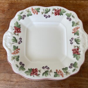 Puede incluir: Un plato cuadrado para servir de cerámica blanca con un borde festoneado. El plato está decorado con un borde de hojas verdes, bayas rojas y negras y un contorno marrón.