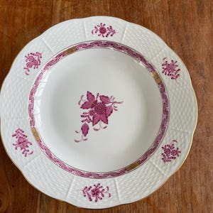 Op de afbeelding: Witte keramische plaat met een geschulpte rand, met een geweven textuur en een decoratieve rand met gouden accenten. De plaat is versierd met roze bloemmotieven, waaronder een centrale cluster en kleinere motieven.