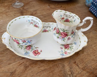 Puede incluir: Un juego de jarra de crema y azucarero de porcelana blanca con un patrón floral y ribete dorado. El juego presenta las palabras "Flowers of the Month" en ambas piezas.