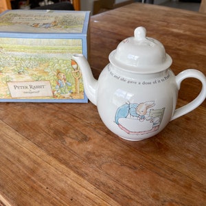 May include: A white teapot featuring a Peter Rabbit design. The teapot has a handle and a spout. The design depicts Peter Rabbit in bed with a cup of tea. The text on the teapot reads "...and she gave a dose of it to Peter".