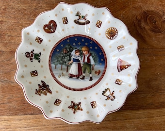 Może przedstawiać: Biały ceramiczny talerz świąteczny z falistym brzegiem. W centrum znajduje się zimowa scena z dwojgiem dzieci spacerujących po śniegu. Talerz jest ozdobiony piernikami, gwiazdami i płatkami śniegu.
