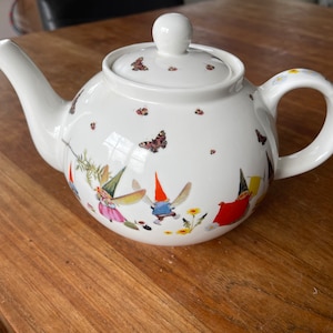 Peut inclure: Théière en céramique blanche avec un design fantaisiste mettant en scène des fées et des gnomes. La théière a un couvercle avec un bouton et une poignée sur le côté. Le design comprend des papillons, des fleurs et d'autres éléments d'inspiration naturelle.