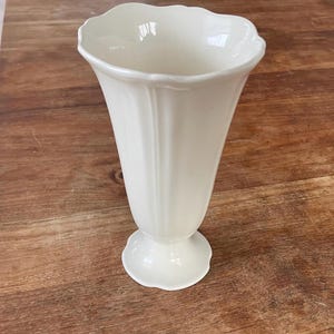 May include: A cream-coloured, fluted vase with a scalloped rim and a pedestal base. The vase has a classic, elegant design, suitable for displaying flowers or as a decorative piece. The vase is approximately 20 cm tall.