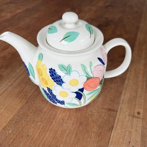 Könnte beinhalten: Weiße Keramik-Teekanne mit floralem Design. Die Teekanne hat einen Deckel mit kleinem Knauf und Henkel. Der Korpus ist mit bunten Blumen in Gelb, Blau, Rosa und Orange verziert.