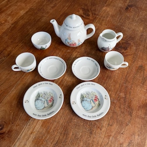 May include: A white ceramic tea set featuring Peter Rabbit illustrations and text. The set includes a teapot, a creamer, two cups, two saucers, and two plates. The plates are inscribed with the text, Once upon a time there were four little Rabbits.