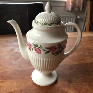 Puede incluir: Cafetera de cerámica blanca con un diseño floral con rosas rosas y hojas verdes. La cafetera tiene una tapa con un pomo en la parte superior y un pico en el lateral. La cafetera está sobre una superficie de madera.