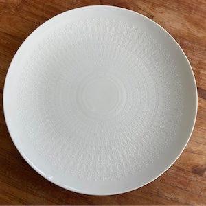May include: White ceramic plate with a raised, embossed design of a circular pattern. The plate has a smooth, glossy finish.