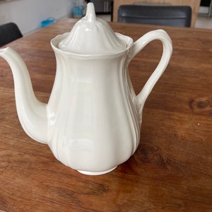 Puede incluir: Una tetera de cerámica blanca con tapa y asa. La tetera tiene un cuerpo liso y redondeado con un pico ligeramente acampanado.
