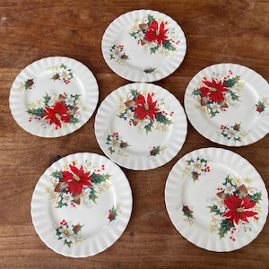 Puede incluir: Seis platos blancos con borde festoneado con un diseño de flor de Pascua navideña. Cada plato presenta una flor de Pascua roja central rodeada de hojas verdes, flores blancas y piñas. Los platos están dispuestos sobre una superficie de madera.