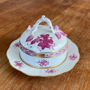 Op de afbeelding: Een witte porseleinen botervloot met een roze bloemenpatroon en een gouden rand. De vloot heeft een deksel met een handvat en een bijpassende voet.