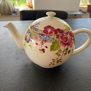 Peut inclure: Une théière blanche avec un motif floral composé de fleurs roses, violettes et vertes. La théière a un couvercle et une anse.