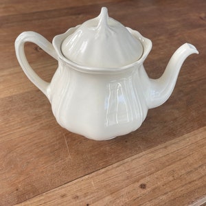 Op de afbeelding: Crèmekleurige keramische theepot met bijpassende deksel. De theepot heeft een gebogen handvat en een tuit. Het deksel is bekroond met een kleine, decoratieve knop. De theepot staat op een houten oppervlak.