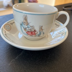 Op de afbeelding: Een wit theeservies met een blauwe rand en een ontwerp van Peter Rabbit en drie konijnen. De tekst "The Tale of Peter Rabbit" staat op de schotel gedrukt.