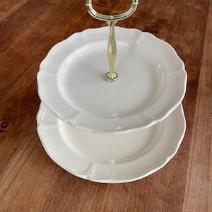 Könnte beinhalten: Ein zweistöckiger Servierständer aus weißem Keramik mit einem goldenen Metallgriff. Der Ständer hat einen gewellten Rand und ist perfekt zum Präsentieren von Desserts oder Vorspeisen.