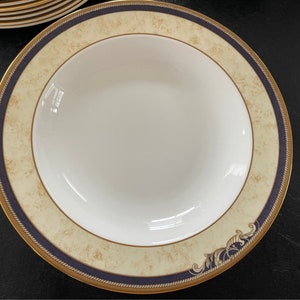 Puede incluir: Un tazón de sopa blanco con un borde dorado y azul. El tazón tiene un fondo beige con un diseño floral dorado y marrón.