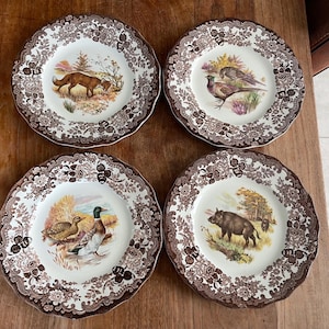 Könnte beinhalten: Vier dekorative Teller mit gewellten Rändern. Jeder Teller zeigt eine zentrale Illustration von Wildtieren, darunter ein Fuchs, ein Fasan, Enten und ein Wildschwein, umgeben von einem braunen Blumenrand. Die Teller sind auf einer Holzoberfläche angeordnet.
