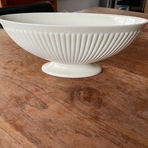 Puede incluir: Un cuenco de cerámica color blanco roto con una abertura ovalada ancha y una textura exterior acanalada y estriada. El cuenco presenta un borde liso y curvado y se apoya sobre una base de pedestal ovalada sólida, con un diseño funcional.