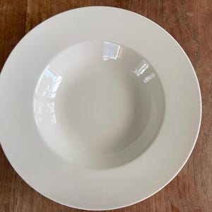 May include: A white ceramic soup bowl with a wide, shallow rim and a smooth, glossy finish.