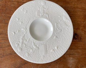 May include: White ceramic plate with an embossed design of stylised figures and foliage. The plate has a central well for serving.