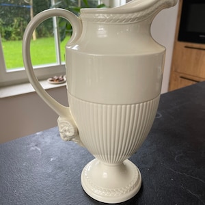Op de afbeelding: Een witte keramische kan met een geribbeld lichaam en een decoratief handvat. De kan heeft een klassiek ontwerp en is perfect voor het serveren van dranken.