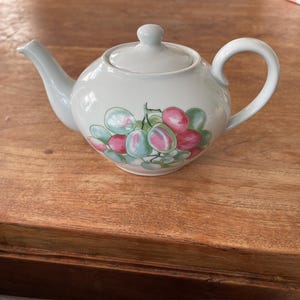 Peut inclure: Une théière en céramique blanche avec un couvercle et une anse. La théière est décorée d'une grappe de raisins roses, verts et bleus. La théière est posée sur une surface en bois.