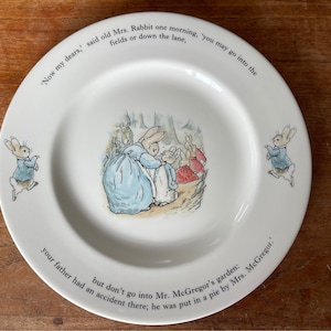 May include: A white ceramic plate featuring a blue and white illustration of Peter Rabbit and his mother. The plate has a quote from the book "The Tale of Peter Rabbit" around the edge: "Now my dears,' said old Mrs. Rabbit one morning, you may go into the fields or down the lane, but don't go into Mr. McGregor's garden: your father had an accident there; he was put in a pie by Mrs. McGregor."
