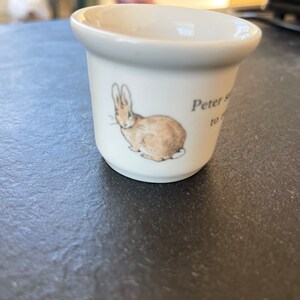 Op de afbeelding: Een witte keramische eierdop met een bruine en bruine illustratie van een konijn. De tekst "Peter says to" staat op de zijkant. De eierdop heeft een uitlopende rand en een cilindrisch lichaam.