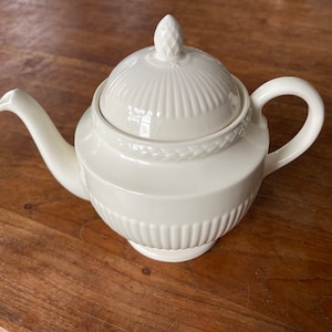 May include: A white ceramic teapot with a lid and a decorative design. The teapot has a rounded body and a spout on one side. The lid has a small knob on top.