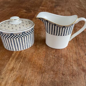 May include: A white ceramic sugar bowl with a lid and a creamer. Both pieces have a navy blue and white striped design with gold trim. The sugar bowl is round, and the creamer has a handle and a spout.