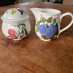 Op de afbeelding: Een witte keramische suikerpot en roomkan set met een bloemenmotief. De suikerpot heeft een deksel en heeft perziken en bladeren. De roomkan heeft pruimen en bladeren.