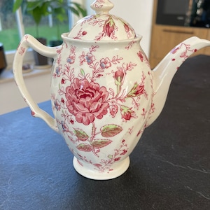 May include: A white ceramic teapot with a floral pattern in shades of pink, green, and blue. The teapot has a lid and a spout.