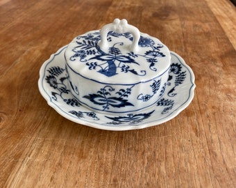 May include: A white porcelain butter dish with a scalloped edge and a matching lid featuring a decorative handle. The dish and lid are decorated with a blue floral pattern, including stylized leaves and blossoms, in a traditional onion-style design.