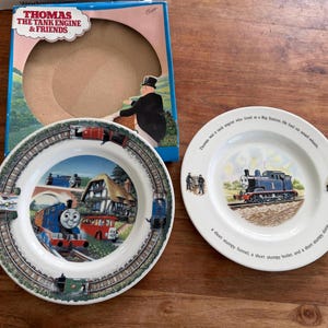 Pode incluir: Dois pratos de cerâmica branca com imagens de Thomas the Tank Engine, acompanhados por uma caixa de cartão azul com a etiqueta Thomas the Tank Engine & Friends. Um prato mostra uma cena de comboio, o outro texto e um comboio azul central.