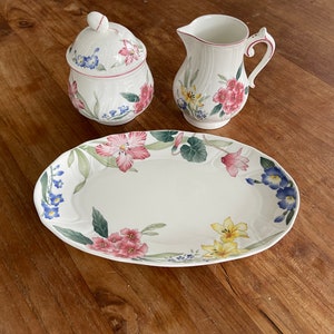 Op de afbeelding: Een witte ovale serveerschaal met een bloemenontwerp met roze, gele, blauwe en groene bloemen. De schaal wordt omringd door een witte suikerpot en roomkan met bijpassende bloemenontwerpen.