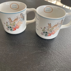 Peut inclure: Deux tasses en céramique blanche avec une bordure bleue et un motif Peter Rabbit. Les tasses présentent un lapin brun courant dans un jardin avec des feuilles vertes et des fleurs. Le texte "Peter Rabbit" est imprimé sur le devant de chaque tasse.