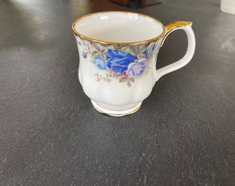 May include: A white teacup with a gold rim and a floral pattern featuring blue and pink roses. The teacup has a handle.