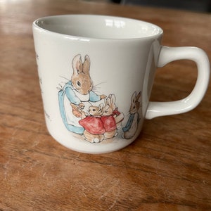 Puede incluir: Taza de cerámica blanca con asa, con una ilustración colorida de Peter Rabbit y otros tres conejos. La ilustración muestra a Peter Rabbit con un vestido azul y sosteniendo una zanahoria. Los otros conejos llevan chaquetas rojas y están de pie detrás de Peter Rabbit.