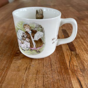 Op de afbeelding: Witte keramische mok met een handvat. De mok heeft een kleurrijke illustratie van een meisje en een konijn uit het boek "Mrs. Tiggy-Winkle" van Beatrix Potter. De tekst "Mrs. Tiggy-Winkle" staat op de mok gedrukt.