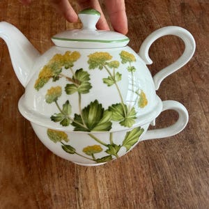 Op de afbeelding: Een witte keramische thee-voor-één set bestaande uit een theepot en een bijpassend theekopje die op elkaar zijn gestapeld. De set is voorzien van een botanisch patroon van gele bloemen en groene bladeren. De theepot heeft een deksel en een handvat.