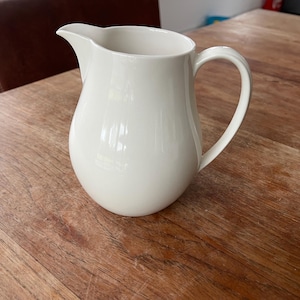 Op de afbeelding: Een witte keramische kan met een gebogen handvat en een schenktuit. De kan staat op een houten tafel. De kan heeft een gladde, glanzende afwerking en een afgeronde body. De handgreep is gebogen. Het is ontworpen voor het serveren van dranken.